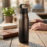 A drink bottle in matte black with a handle, placed on a wooden table beside a notebook and a plant.
