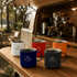 A set of five reusable coffee cups in white, red, orange, black, and blue, featuring a logo, placed on a wooden counter.