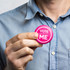 A round pink button badge held by a person wearing a blue collared shirt. The badge features a logo.