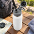A drink bottle with a stainless steel body in white, featuring a grey lid, placed on a wooden surface with outdoor items.