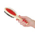 A pet grooming brush with a cream handle, red rubber pad, and black bristles, held in a hand with red nails.