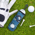 A golf ball holder in blue with graphics, accompanied by a golf glove, golf ball, and tee on green grass.