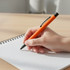 An orange metal pen held in a hand above a graph notebook on a wooden surface.