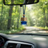 A car reed diffuser in blue glass with a wooden top, hanging from a rearview mirror amidst a forest backdrop.