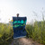 A smiling man in glasses and a cap holds up a puffy camping quilt printed with a teal and green aurora borealis design over dark mountains, standing on a trail surrounded by tall grass and wildflowers.