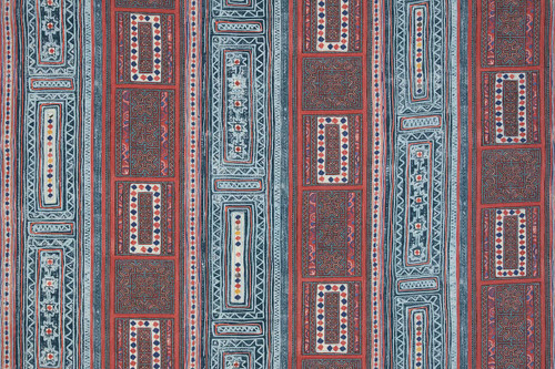 A vibrant textile with vertical stripes and geometric patterns featuring diamonds, florals, and intricate borders in red, blue, white, and yellow.
