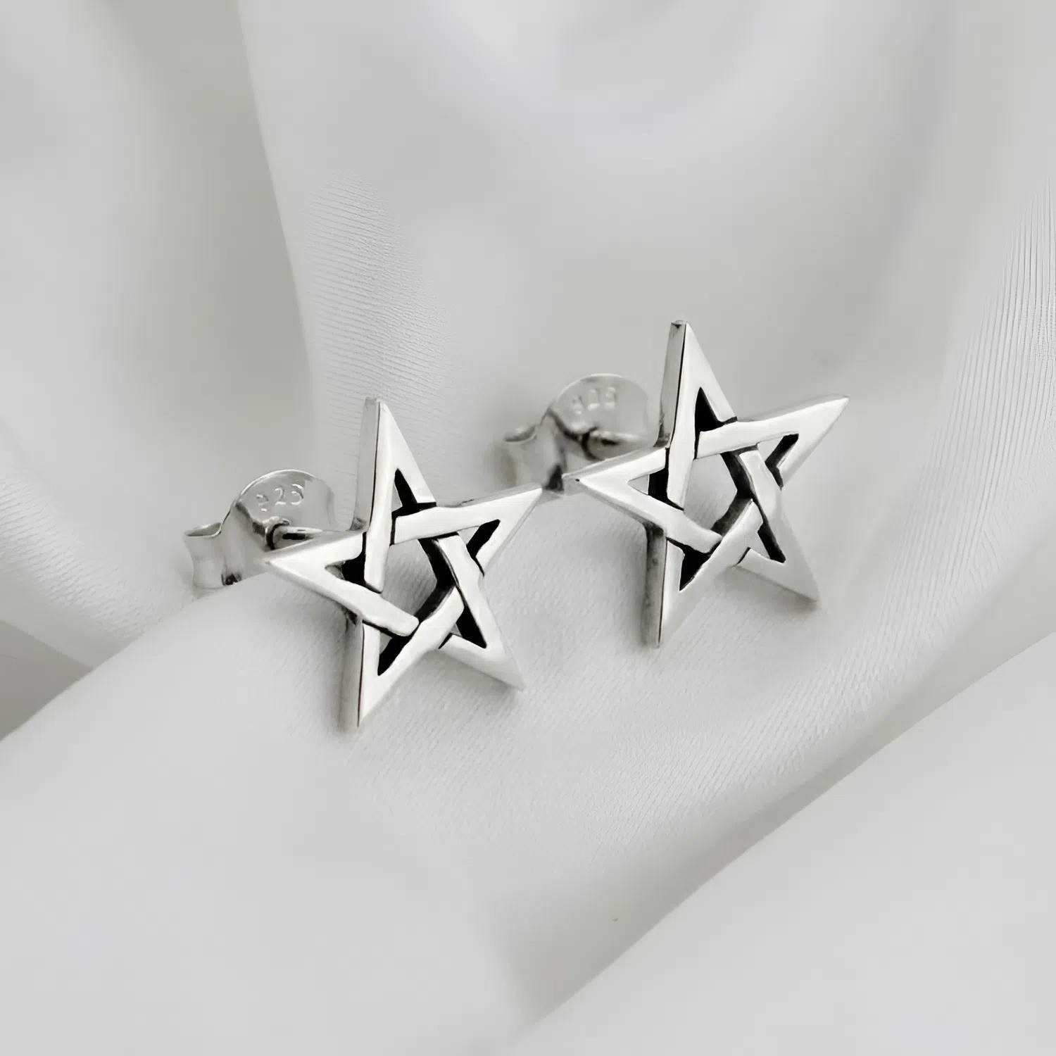 Pentagram post earrings capturing a sleek, woven star aesthetic, displayed gracefully on folded, smooth white silk fabric