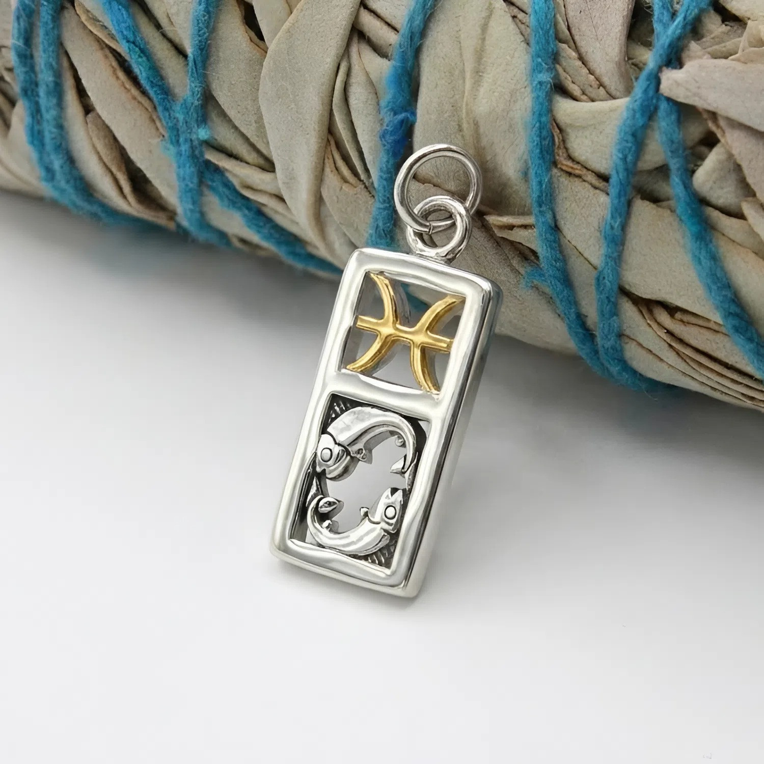 Artistic shot of a Pisces pendant at our pagan jewelry store resting against a bundle of dried sage bound with blue string, highlighting its spiritual theme