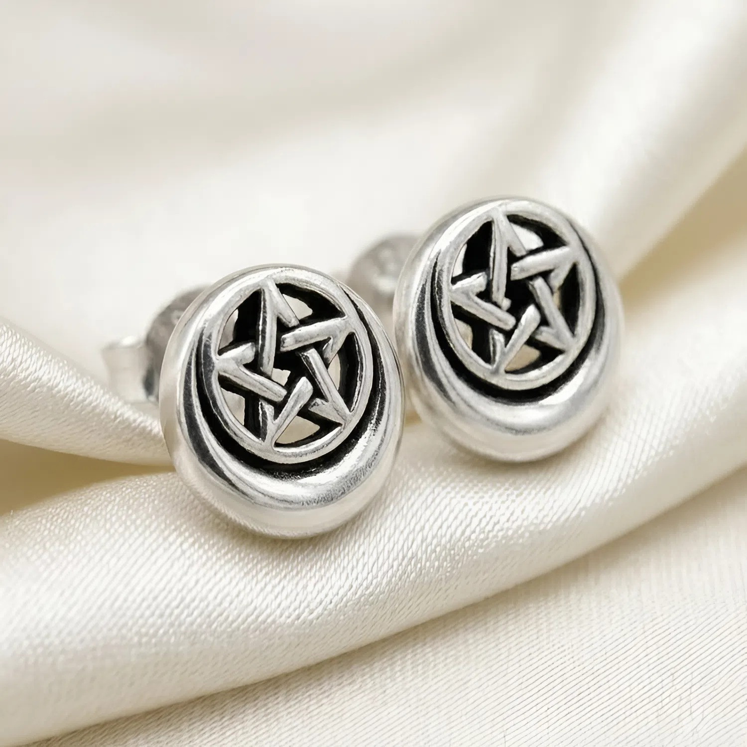 Moon pentacle stud earrings resting on a piece of shimmering white silk fabric