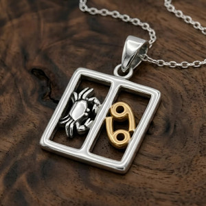 Cancer pendant zodiac jewelry, displaying a two-tone silver and gold horoscope design resting elegantly on a warm, dark wood grain surface