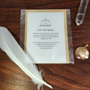 A flat lay image displays a curated collection of witchcraft supplies on a dark wooden surface. At the center is a packaged product card for spell parchment paper, with the title "Craft Your Reality." Surrounding the card are a large white feather, a clear quartz crystal point, and a detailed gold medallion, showcasing essential tools for any ritual. This arrangement of occult store supplies highlights key witchy items and pagan supplies for your craft.