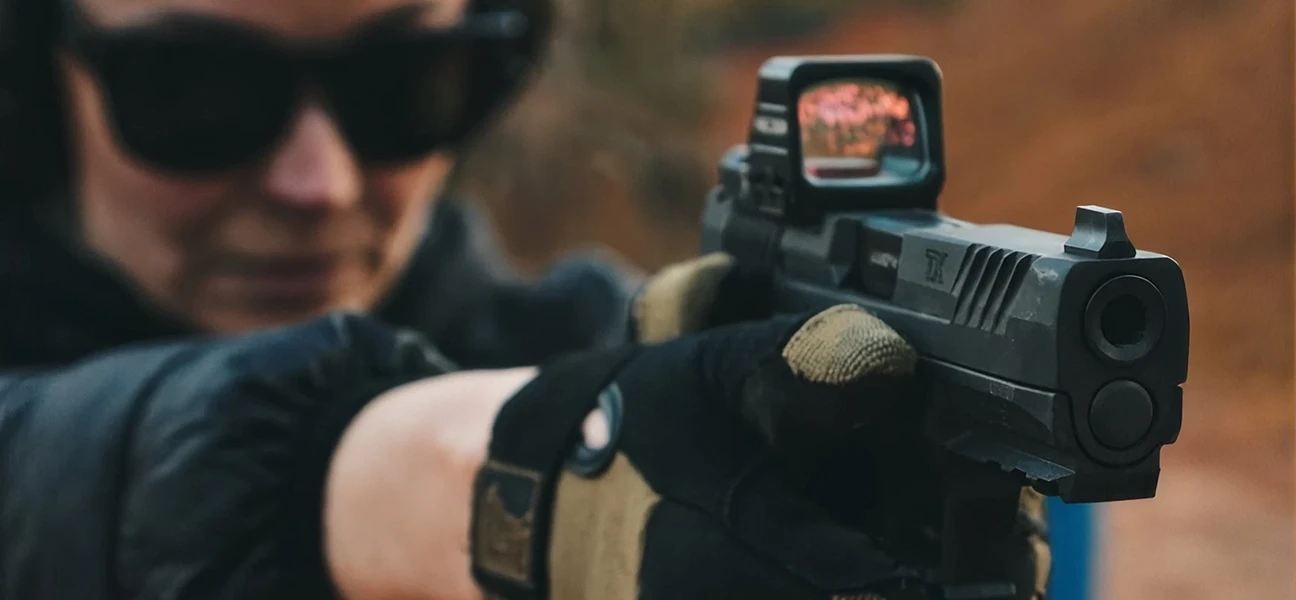 Person aiming a handgun equipped with a red dot sight at an outdoor shooting range