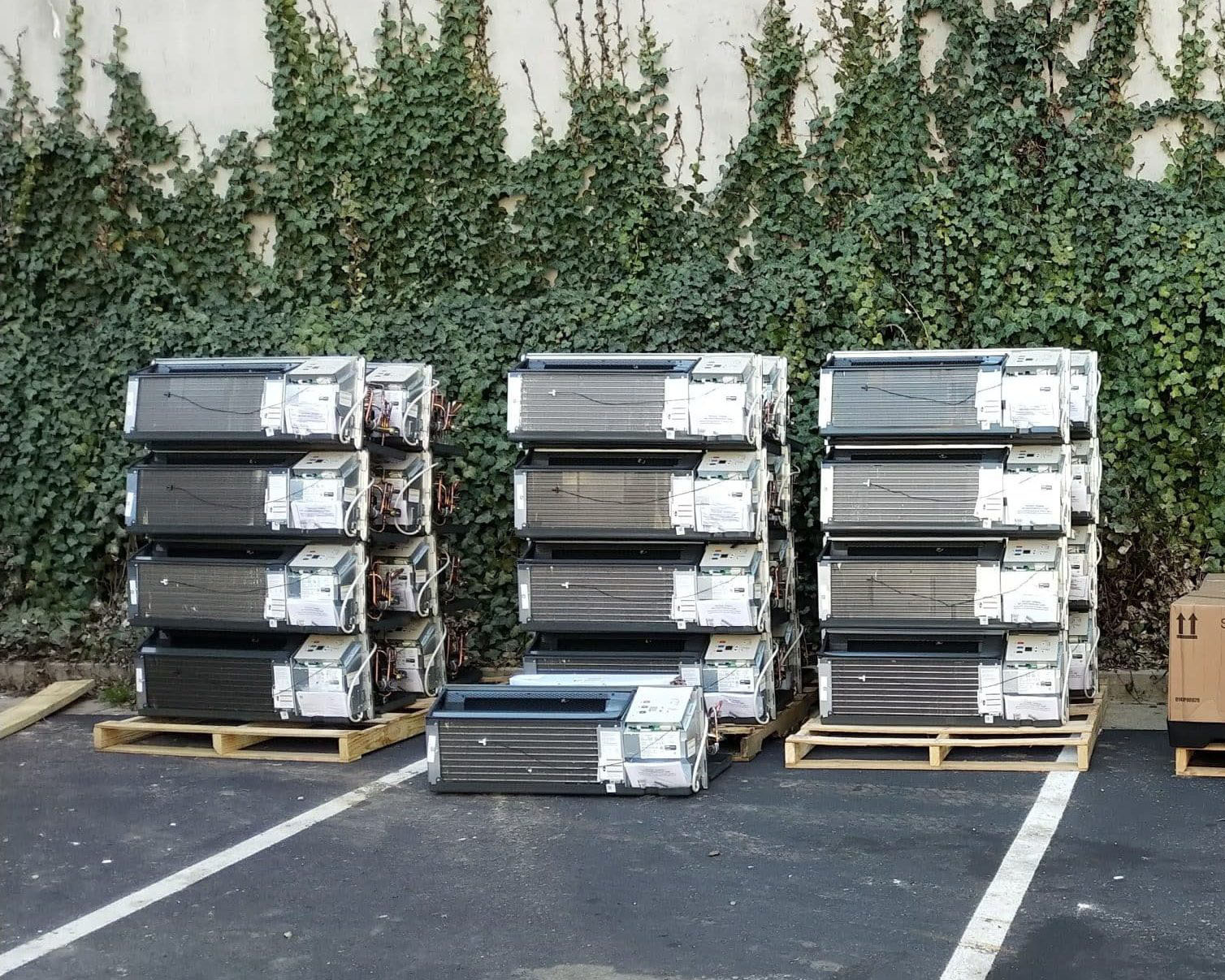 Stacked pallets of used PTAC (Packaged Terminal Air Conditioner) units awaiting proper disposal, lined up against a wall covered in climbing ivy.