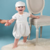 OONA Infant Baby Boy Baptism Romper, a cute baby boy wearing a white baptism romper and matching cap, holding an adult's hand, standing on a fluffy rug next to a vintage-style chair against a blue background.