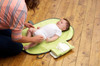 JJOVCE Portable Folding Nappy Changing Pad, a baby lying on a green changing pad with a caregiver changing the baby's diaper on a wooden floor.