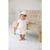 OONA 2 PCS Baptism White Satin Suit with Hat, adorable baby girl in white satin baptism outfit with matching hat, standing next to a beige tufted sofa in a bright, cozy room.