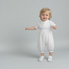 Minnie Me Azaiah Christening Romper, a happy toddler boy wearing a white christening romper with blue embroidery, standing on a plain light grey background.