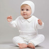 Minnie Me Noah Christening Romper, adorable baby wearing a white christening romper and hat, sitting barefoot, showcasing the detailed embroidery and soft fabric of the baby outfit.