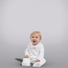 Minnie Me Noah Christening Romper, adorable baby girl wearing a white christening romper with embroidered cross design, sitting on the floor with happy expression.