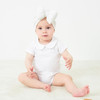 Minnie Me Cotton Romper Neutral, adorable baby girl sitting on a white surface wearing a white romper with a Peter Pan collar and a white headwrap.