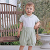 Minnie Me Elie Formal Set with Short, toddler girl wearing a stylish white and green short-sleeved romper with a Peter Pan collar, standing outdoors near a wooden bench.
