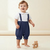 Minnie Me Eric Baby Boy Christening Romper Set, adorable young boy wearing a white and navy blue romper with suspenders, standing barefoot on a light-colored background.