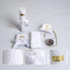 Minnie Me Elegant White Baptism Set, includes a decorated candle with lace and ribbon, a white embroidered towel, a white box with ribbon, and a seashell with a rosary and pearl necklace.
