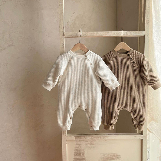 Generic Knitted Round Neck Bodysuit, two baby bodysuits hanging on wooden hangers on a rustic shelf, one in cream color and the other in beige, ideal for infants and toddlers, made of soft knitted fabric with long sleeves and round neck design.