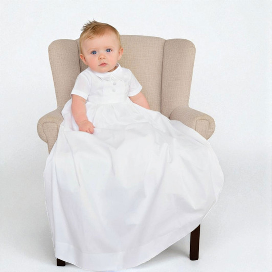 Minnie Me Caleb Boys Christening Gown, a young child sitting in a beige armchair wearing a white christening gown with short sleeves and a full-length skirt.