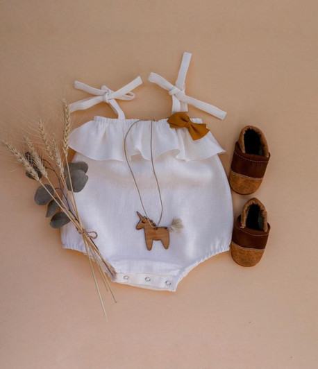 OONA White Bowknot Sleeveless Romper, sleeveless baby romper with bowknot straps and ruffled top, paired with brown baby shoes and wooden unicorn necklace, on a beige background with dried flowers.