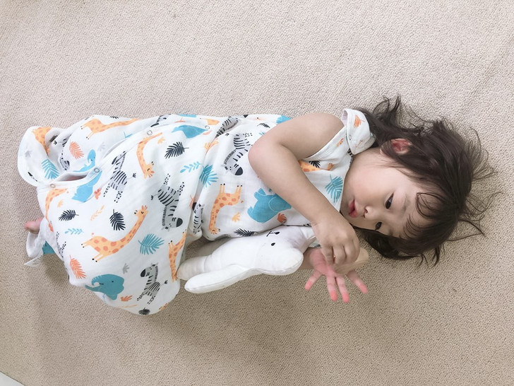 Karitree Four Layer Jungle Sleeveless Sleeping Bag, baby girl lying on a beige carpet with a colorful jungle animal print, holding a plush toy.
