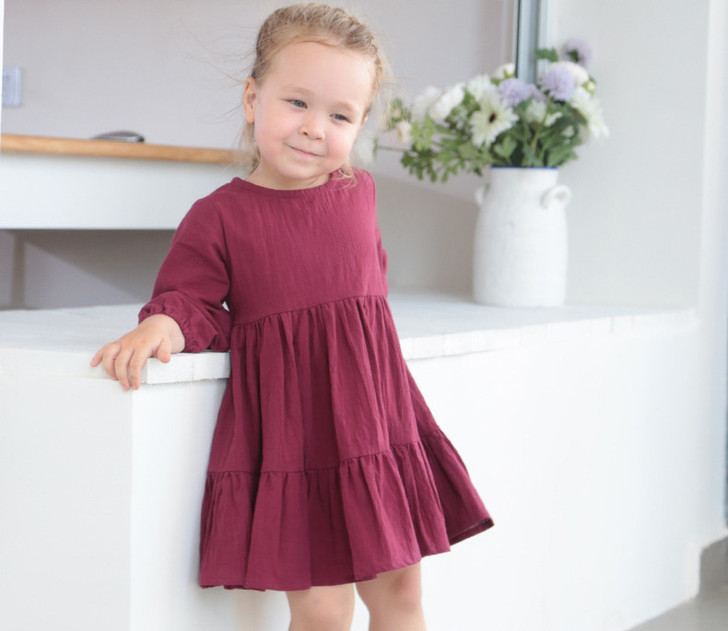 Generic Long Sleeve Linen Solid Colour Dress, A young girl wearing a long sleeve maroon linen dress with a tiered skirt, standing indoors near a white counter with a vase of flowers in the background.