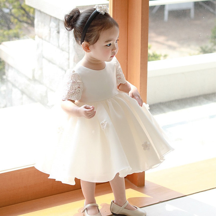 OONA Daisy Dress Ivory White, adorable young girl wearing a white lace and satin dress with floral details and a headband, standing near a window.