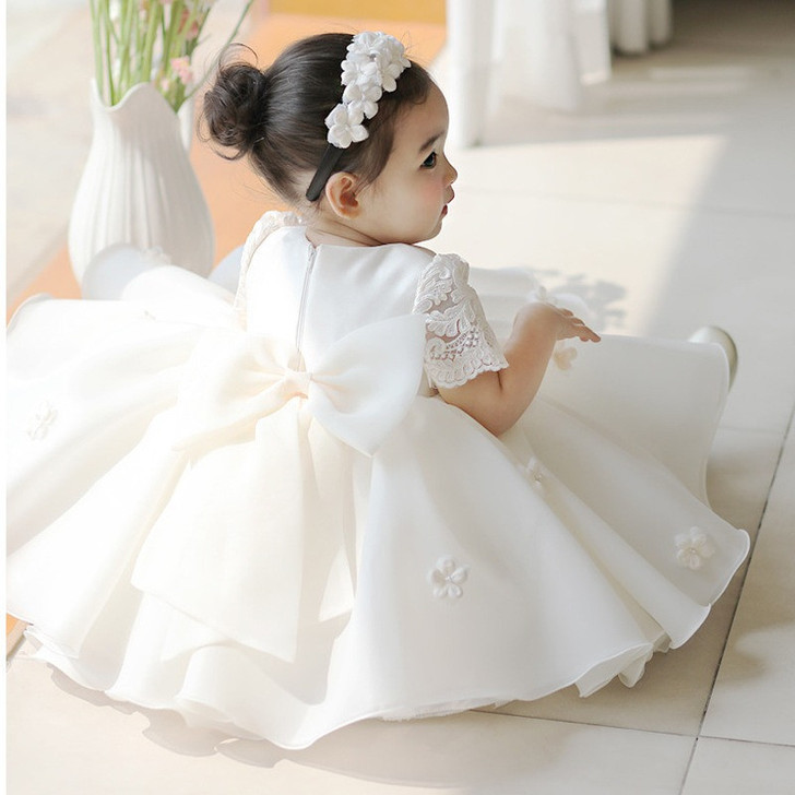 OONA Daisy Dress Ivory White, a beautiful young girl wearing a white lace dress with floral embroidery, a large bow on the back, and a floral headband, sitting on the floor.