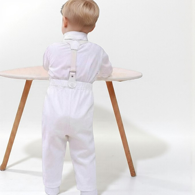 Minnie Me Nicolas Suspender Set, young child standing with back facing, wearing white suspender outfit and shirt, in front of a light-colored desk.