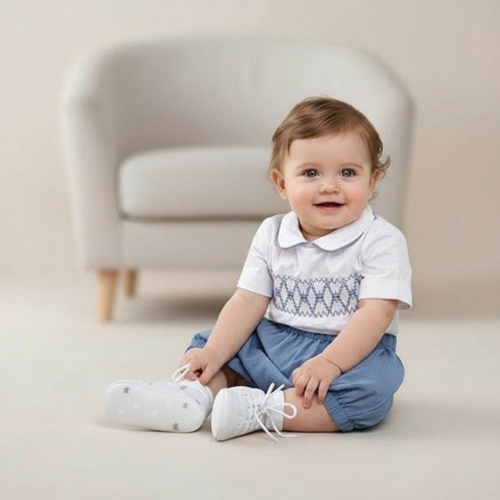 Minnie Me Mateo Formal Short Set, a toddler boy wearing a white shirt with blue embroidery, blue shorts, white sneakers with star patterns, sitting on the floor in front of a white chair.