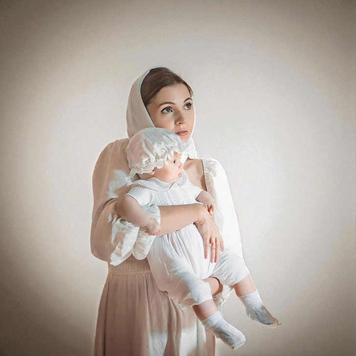 Minnie Me Mike White Baptism Romper, a young woman holding a baby girl dressed in a white baptism romper and bonnet, with both wearing matching white outfits.