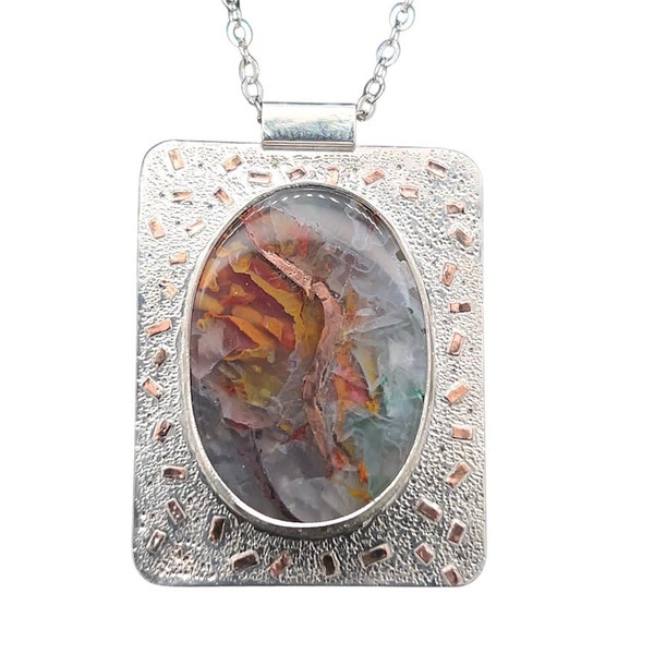 Chrysocolla with Copper Pendant. I cut this large stone from a rare specimen of chrysocolla in quartz from Indonesia. It is rare because of the huge internal crystals of metallic copper that are deposited naturally into the stone. This one has a unique lighting bolt symbol like Happy Potter that runs vertically through the stone. Set with fine silver bezel and copper accents with hand textured and diamond textured imprints that create a sparkle. Includes 18”chain and clasp. 
1 ½ x 1 ¼ inches.