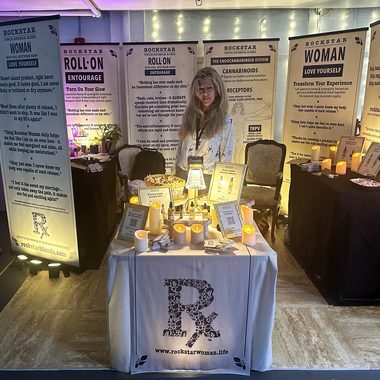 Heather Reilly Hiemstra, founder of Rockstar Blends, stands behind her candlelit botanical apothecary table—product trays and an "R" logo cloth reading rockstarwoman.life—framed by banners explaining the endocannabinoid system and the Rockstar Woman and Roll-On topicals at the Cannadelic Night Market.