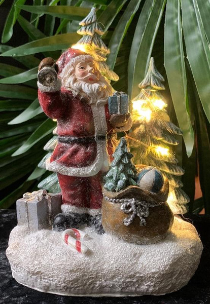 Santa holding a bell and a gift in front of a tree with LED lights