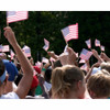 A crowd of people waving 4" x 6” American stick flags. The flag has 50 white stars on a blue canton & 13 alternating red & white stripes.