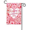 A Valentine's themed garden banner with hues of carnation pink, rose red, and bright white, hangs on a black garden flag pole. In the center of the garden flag is a white heart framed in light pink with cherry red script  spelling 'happy valentine's day.'  The central heart is surrounded and framed with a collage of doily like hearts and flowers.