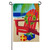 A Coastal Christmas Adirondack Applique Garden Flag depicting an Adirondack chair with a winter wreath on a beach next to some wrapped presents.