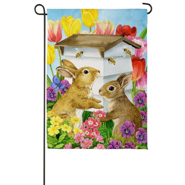 A Bunny Beehive Suede Garden Flag depicting two rabbits in front of a white beehive, surrounded by flowers.