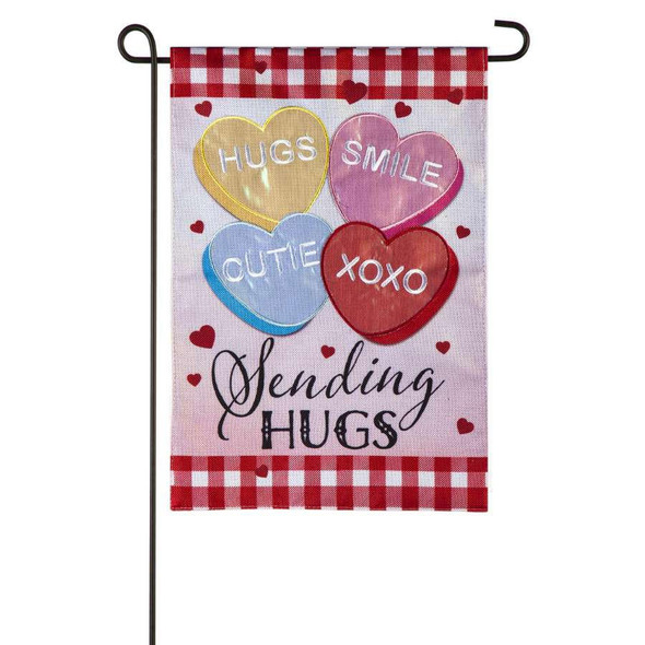 A pink garden flag featuring four differently-colored heart candies imprinted with the words "hugs", "smile", "cutie", and "XOXO". Below the candies the text "sending hugs" is written.