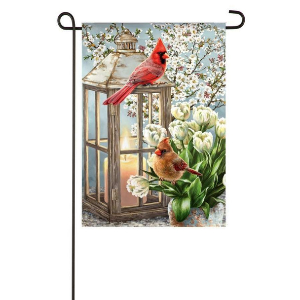 A scenic decorative garden flag with cardinals perch atop a lantern surrounded by dogwood blossoms. The garden flag is shown on a garden flagpole.
