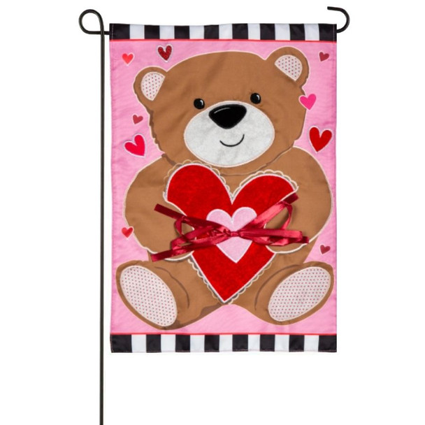 A pink garden flag depicting a brown bear holding a red heart with a pink bow.