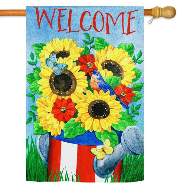 Beautiful bouquet of five sunflowers and three red flowers all sitting inside of an American flag painted watering can. The can sits on top of grass and the background is a blue sky. On the top is red text that reads "welcome" in all caps.