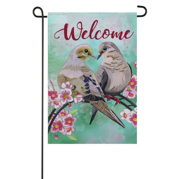 A turquoise garden flag featuring the text ‘Welcome’ along the top, under which two mourning doves are perched on a branch, surrounded by pink flowers.