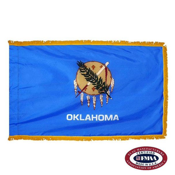The indoor Oklahoma flag has 3 edges of gold fringe around the blue field. In the center is a shield with feathers, a calumet, & an olive branch. FMAA Certified Made in America seal.